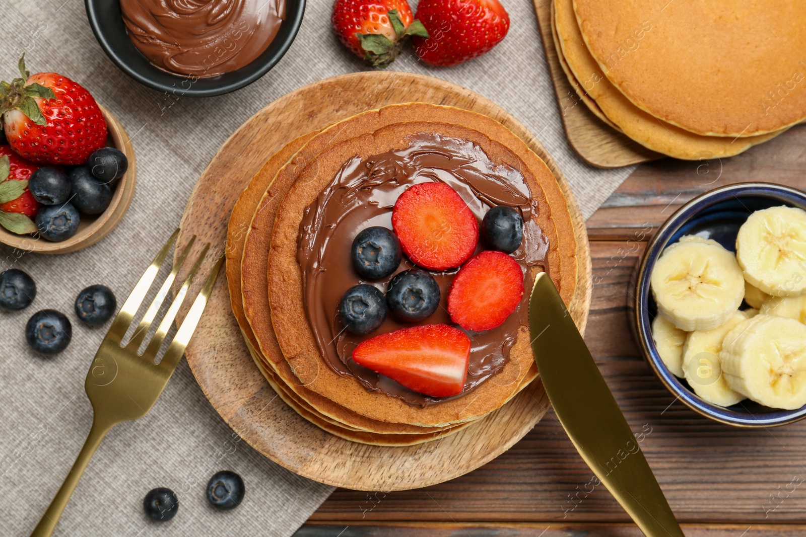 Photo of Tasty pancakes with chocolate paste, berries and banana served on wooden table, flat lay