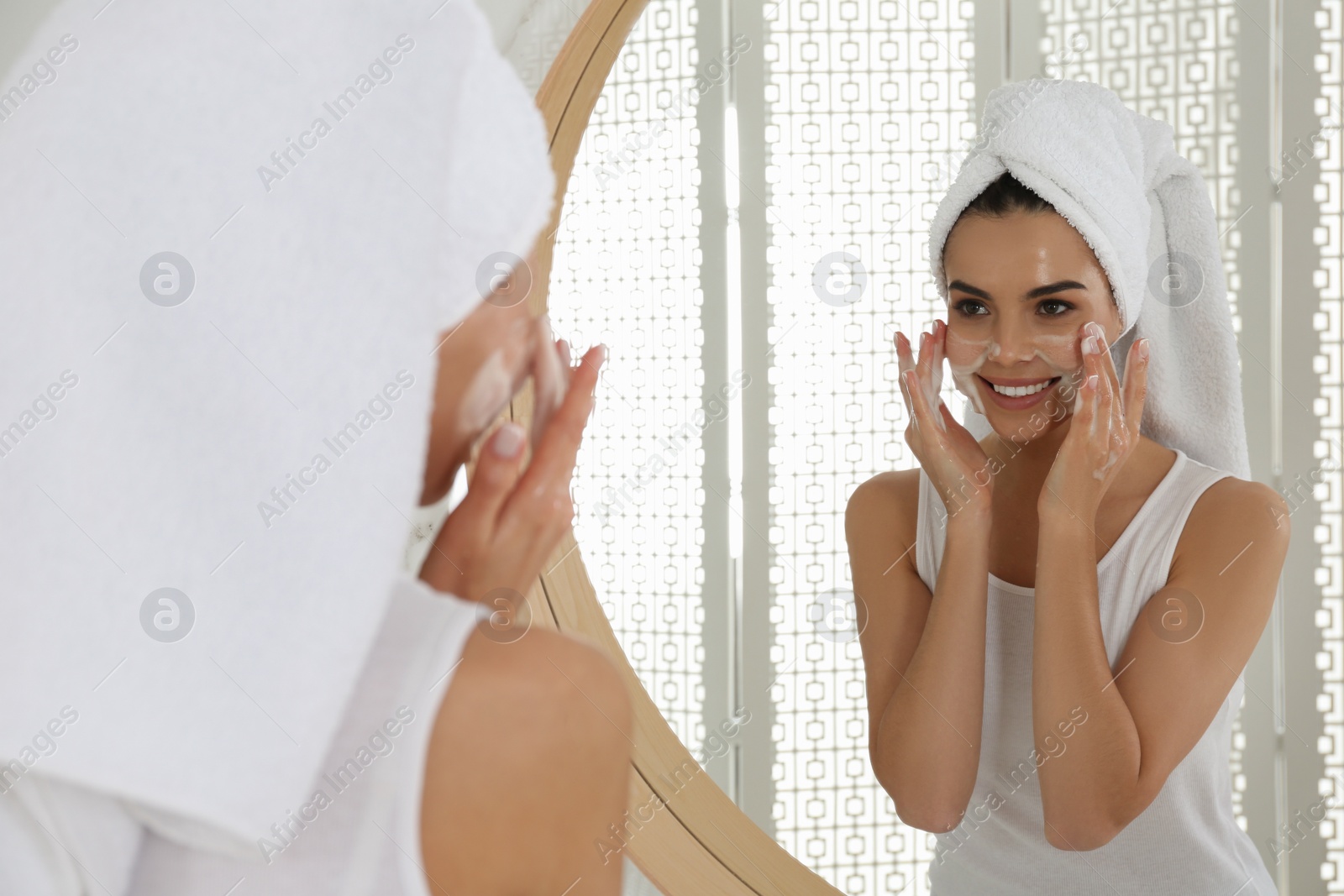 Photo of Happy young woman applying cleansing foam onto face near mirror in bathroom