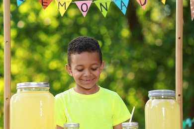 Cute little African-American boy with money at lemonade stand in park. Summer refreshing natural drink