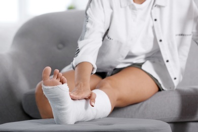 Photo of Young woman with broken leg in cast sitting on sofa at home