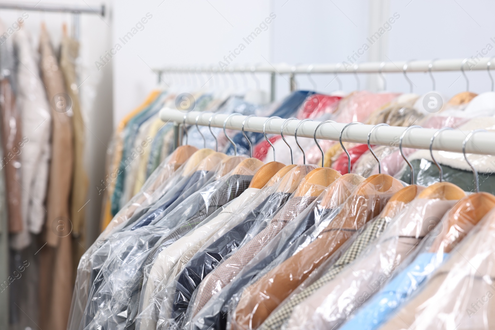 Photo of Dry-cleaning service. Many different clothes in plastic bags hanging on rack indoors, closeup