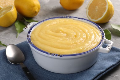 Delicious lemon curd in bowl, fresh citrus fruits, green leaves and spoon on grey table, closeup