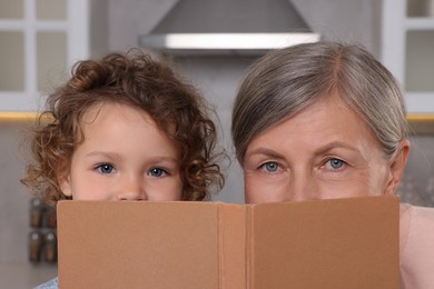 Photo of Cute little girl and her granny with recipe book in kitchen, closeup
