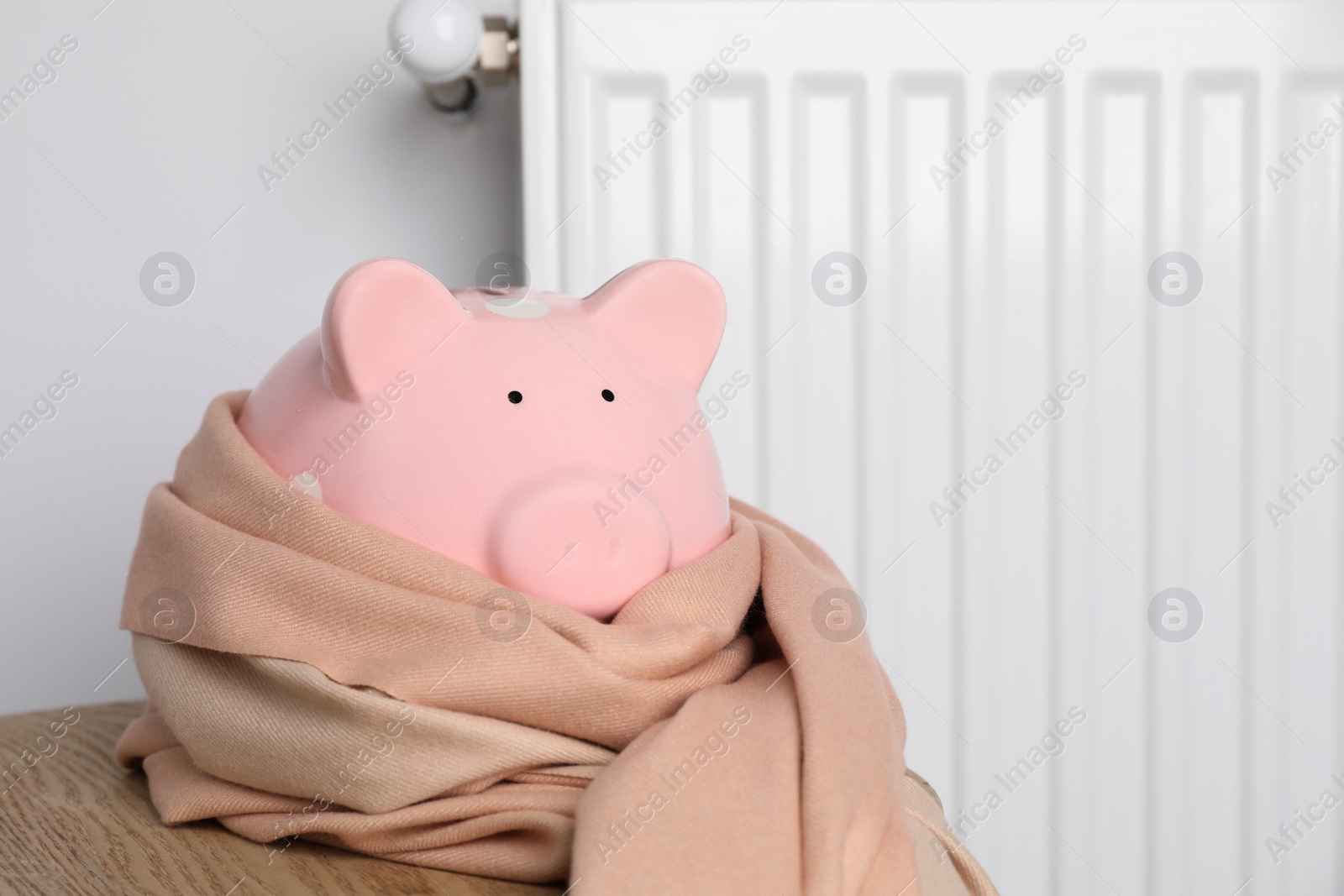 Photo of Piggy bank wrapped in scarf on wooden table near heating radiator. Space for text