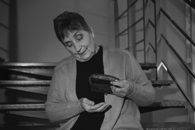 Photo of Poor senior woman with empty wallet sitting on stairs indoors. Black and white effect