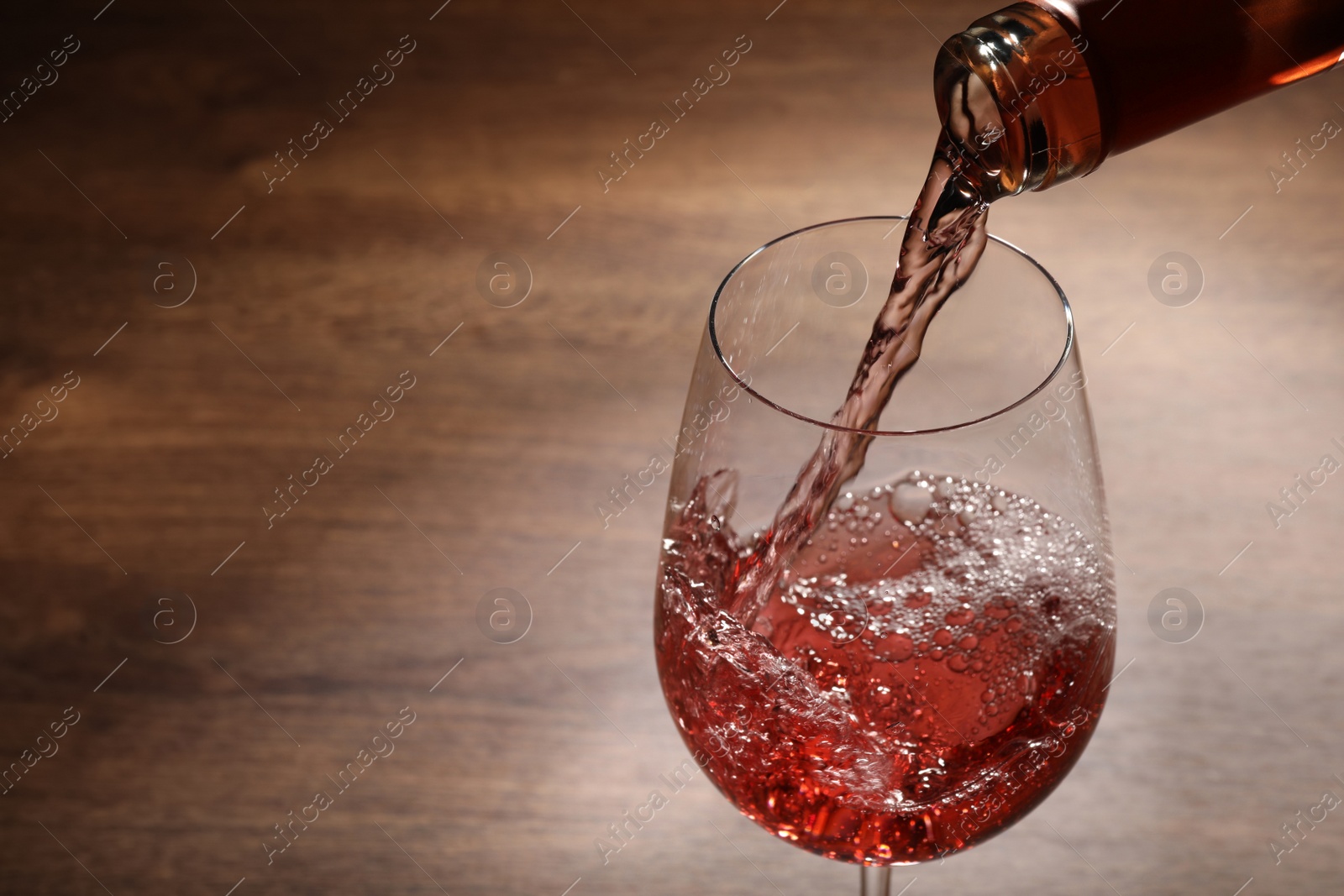 Photo of Pouring rose wine from bottle into glass on wooden table, closeup. Space for text