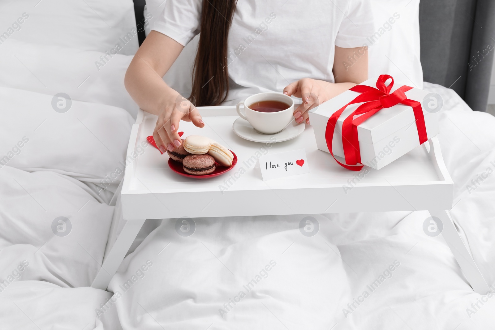 Photo of Tasty breakfast served in bed. Woman with tea, macarons, gift box and I Love You card at home, closeup