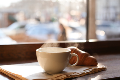 Photo of Delicious morning coffee, newspaper and croissant near window, indoors