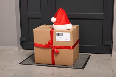 Photo of Christmas gift box and Santa hat near door indoors. Sending present by mail