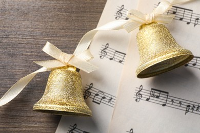 Photo of Golden shiny bells with bows and music sheets on wooden table, flat lay. Christmas decoration