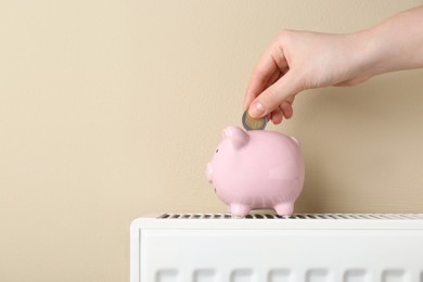 Photo of Woman putting coin into piggy bank on heating radiator against beige background, closeup. Space for text
