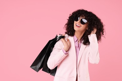 Happy young woman with shopping bags and stylish sunglasses on pink background