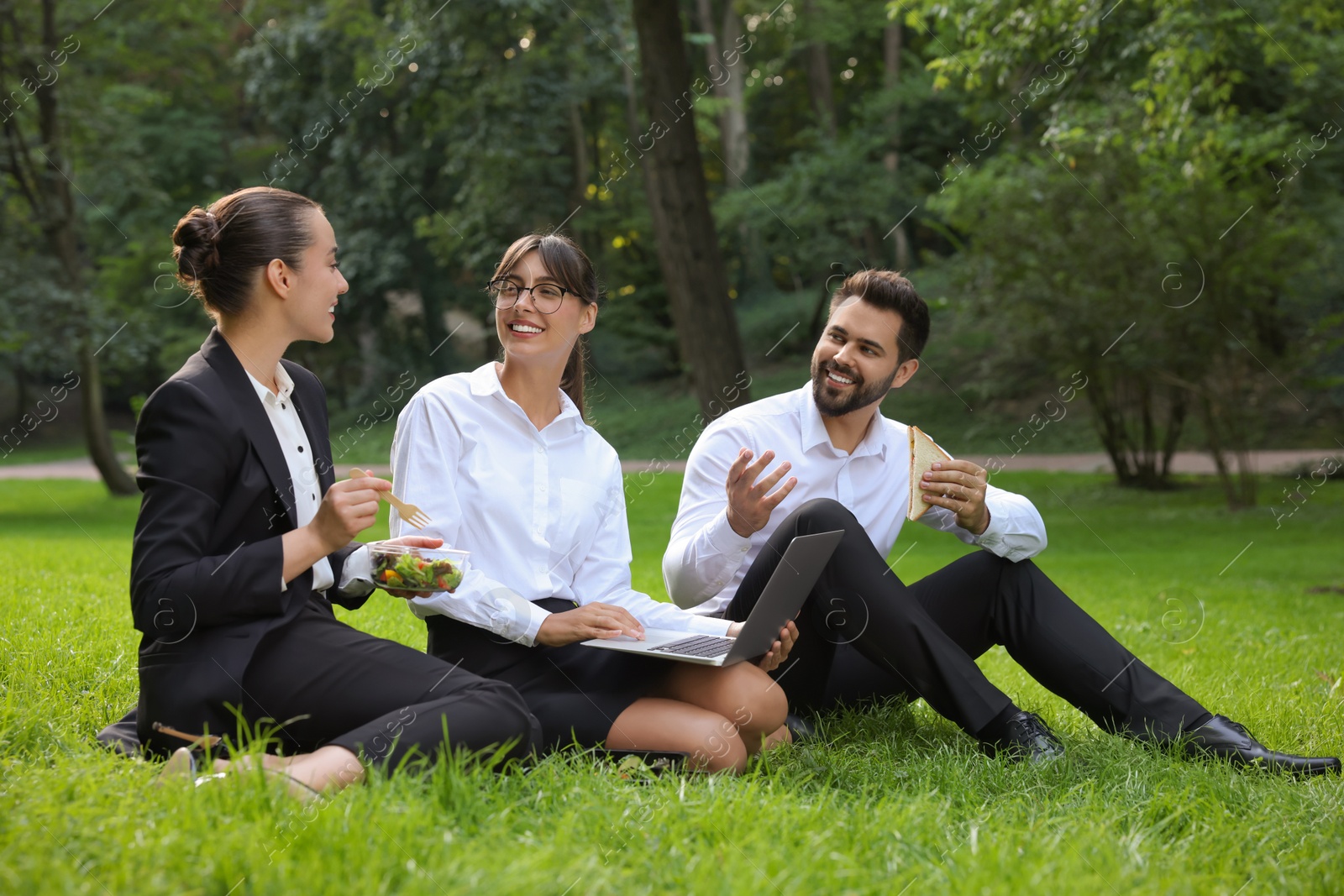 Photo of Happy colleagues with laptop having business lunch on green grass in park