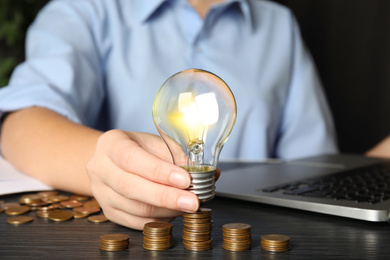 Photo of Woman with light bulb and coins at black wooden table, closeup. Power saving
