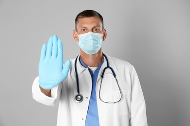 Photo of Doctor in protective mask showing stop gesture on grey background. Prevent spreading of coronavirus