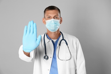 Doctor in protective mask showing stop gesture on grey background. Prevent spreading of coronavirus