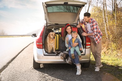 Parents, their daughter and dog sitting in car trunk outdoors. Family traveling with pet