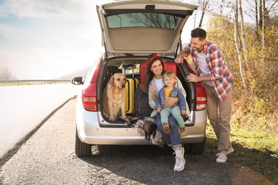 Photo of Parents, their daughter and dog sitting in car trunk outdoors. Family traveling with pet