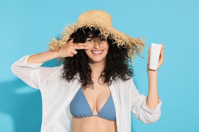 Photo of Beautiful young woman in straw hat with tube of sunscreen on light blue background