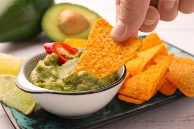 Photo of Woman dipping delicious nachos chip into guacamole at white wooden table, closeup