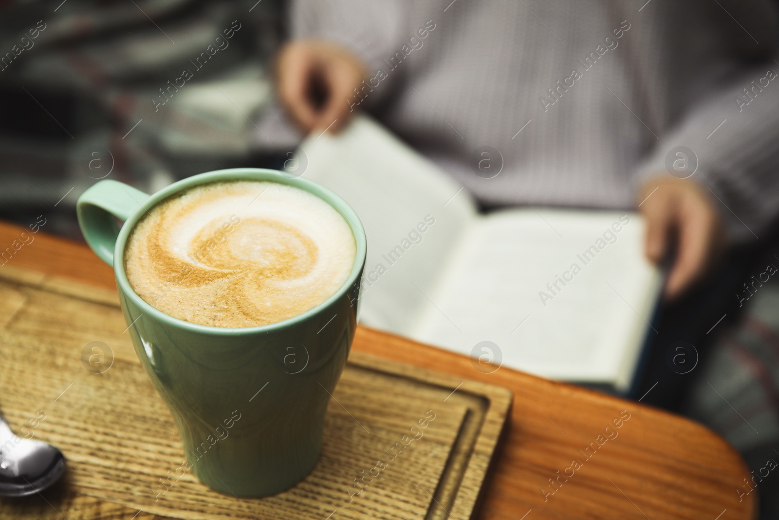 Photo of Woman reading book at table, focus on cup of coffee