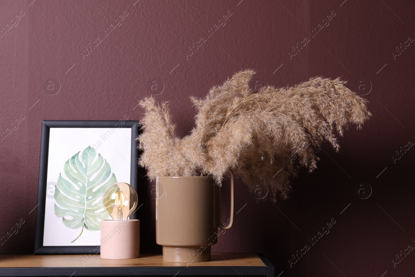 Photo of Ceramic vase with fluffy dry plants, painting and modern night lamp on wooden table near brown wall
