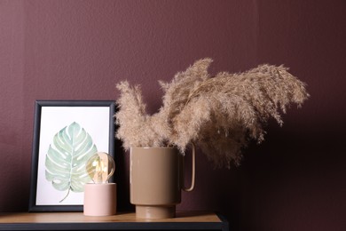 Ceramic vase with fluffy dry plants, painting and modern night lamp on wooden table near brown wall