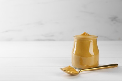 Photo of Spoon and glass jar of tasty mustard sauce on white wooden table. Space for text