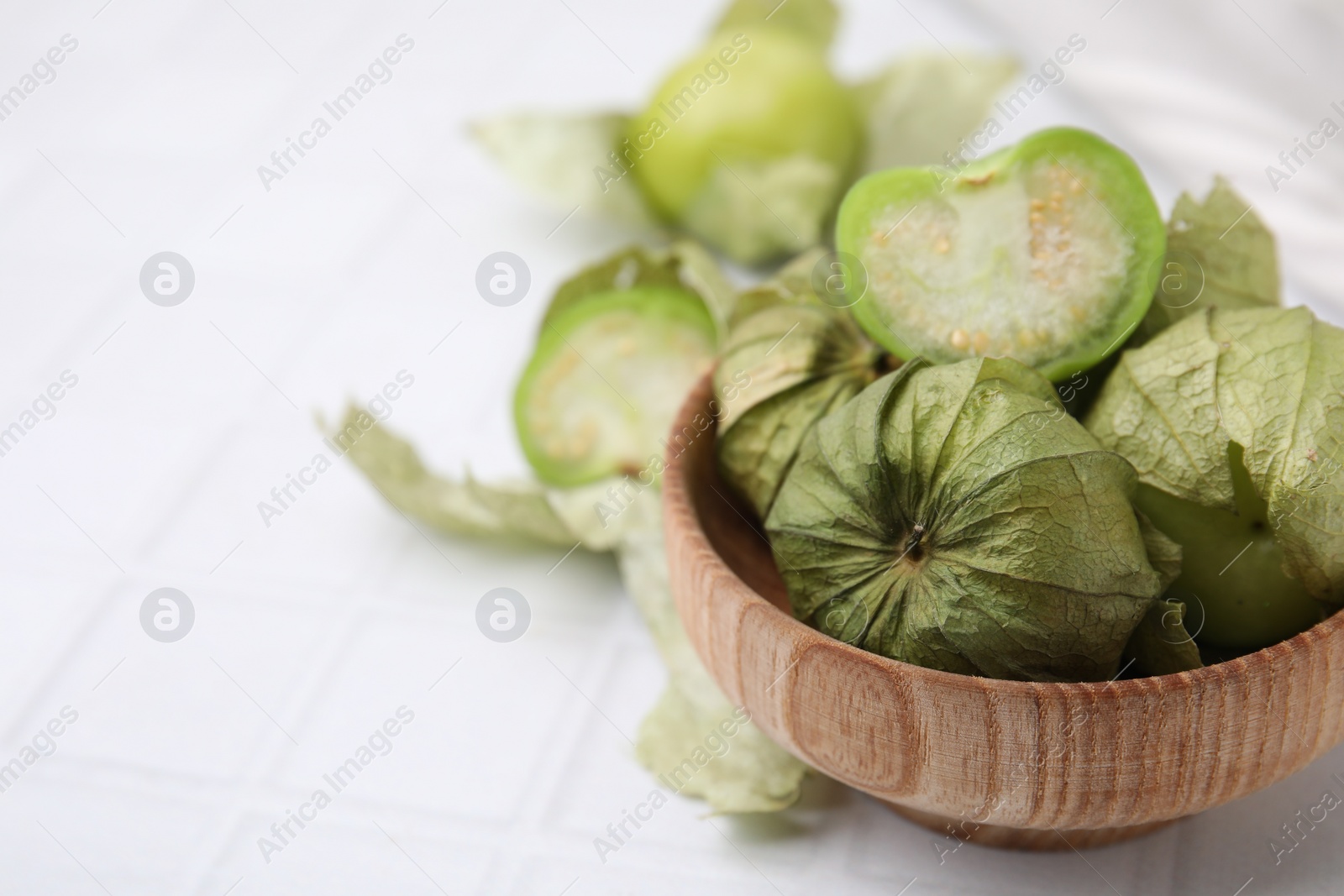 Photo of Fresh green tomatillos with husk in bowl on white tiled table, closeup. Space for text