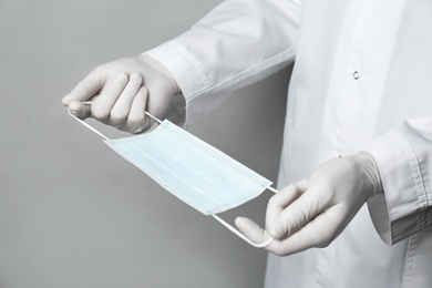 Doctor in medical gloves holding protective mask on light grey background, closeup