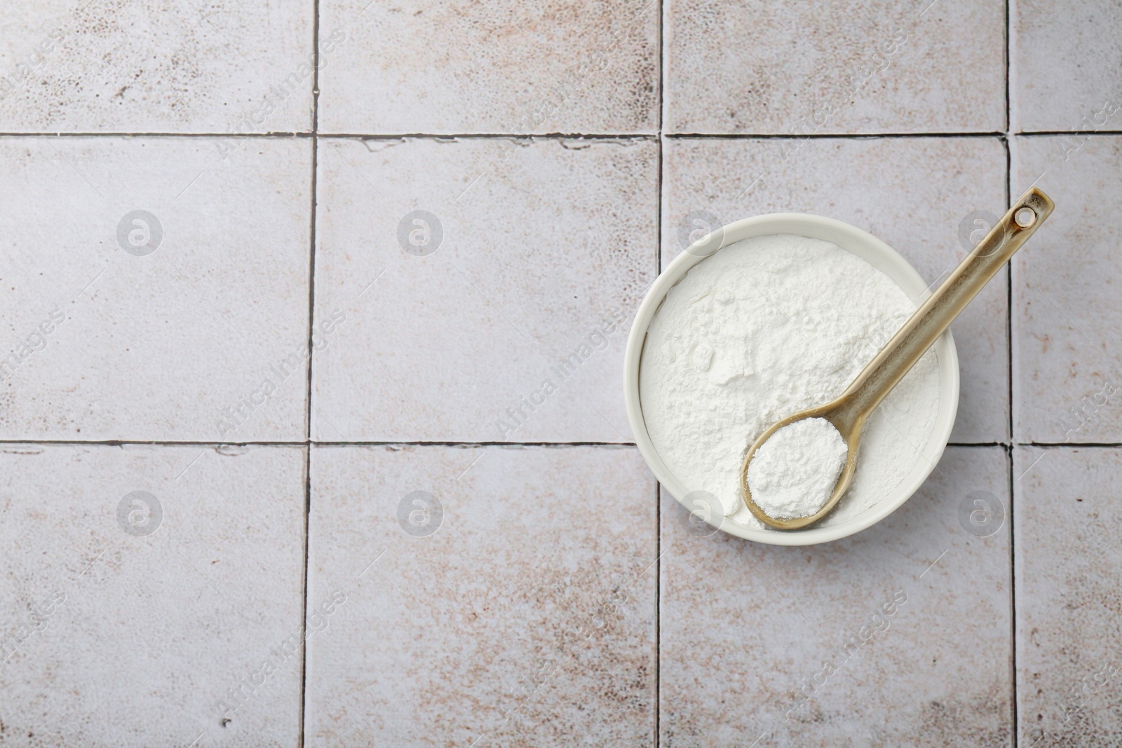 Photo of Baking powder in bowl and spoon on light tiled table, top view. Space for text