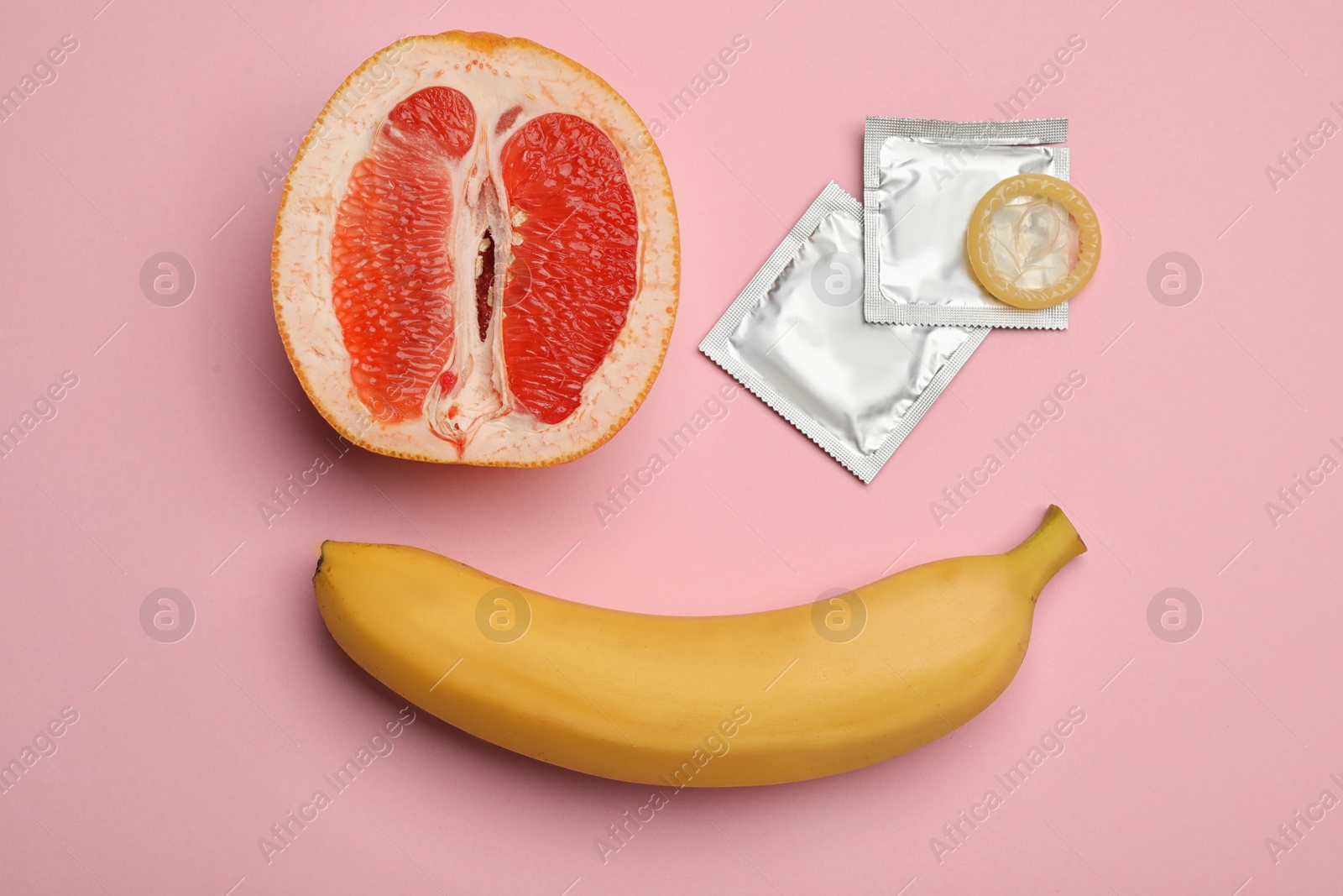 Photo of Condoms with banana and cut grapefruit on pink background, flat lay. Safe sex