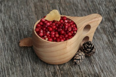 Photo of Cup with tasty ripe lingonberries and cones on wooden surface