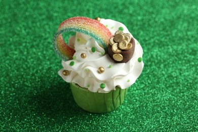 Photo of St. Patrick's day party. Tasty cupcake with sour rainbow belt and pot of gold toppers on shiny green surface, closeup