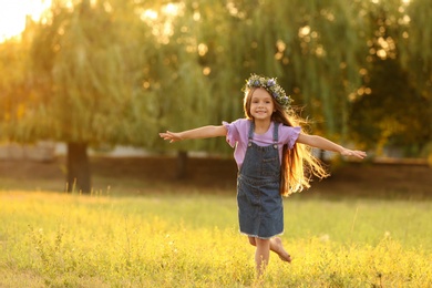 Photo of Cute little girl wearing flower wreath outdoors at sunset, space for text. Child spending time in nature