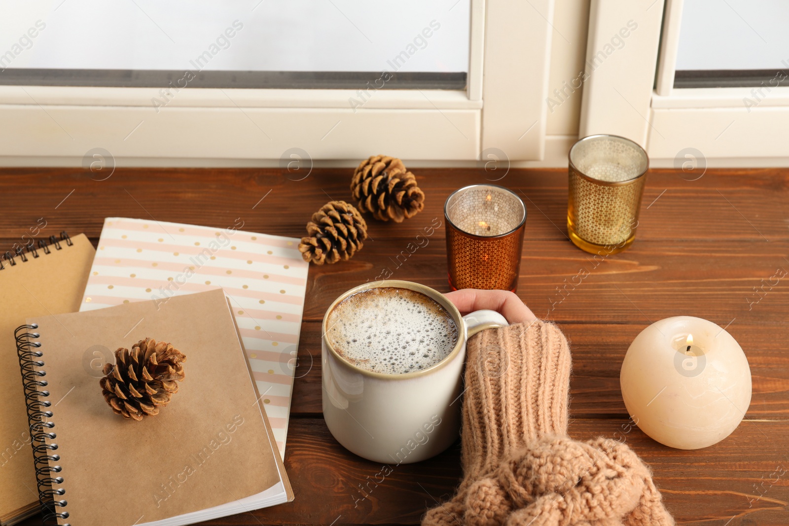 Photo of Woman with cup of coffee and notebooks at window indoors, closeup. Winter drink