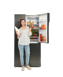 Young woman with milk and pickles near open refrigerator on white background