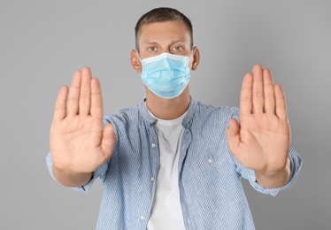 Man in protective mask showing stop gesture on grey background. Prevent spreading of coronavirus