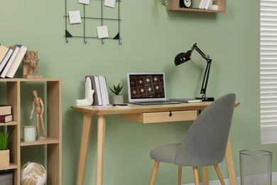 Photo of Workplace with modern laptop on wooden desk, bookcase and cosy chair at home