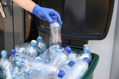 Photo of Woman in gloves putting used plastic bottle into trash bin outdoors, closeup. Recycling problem