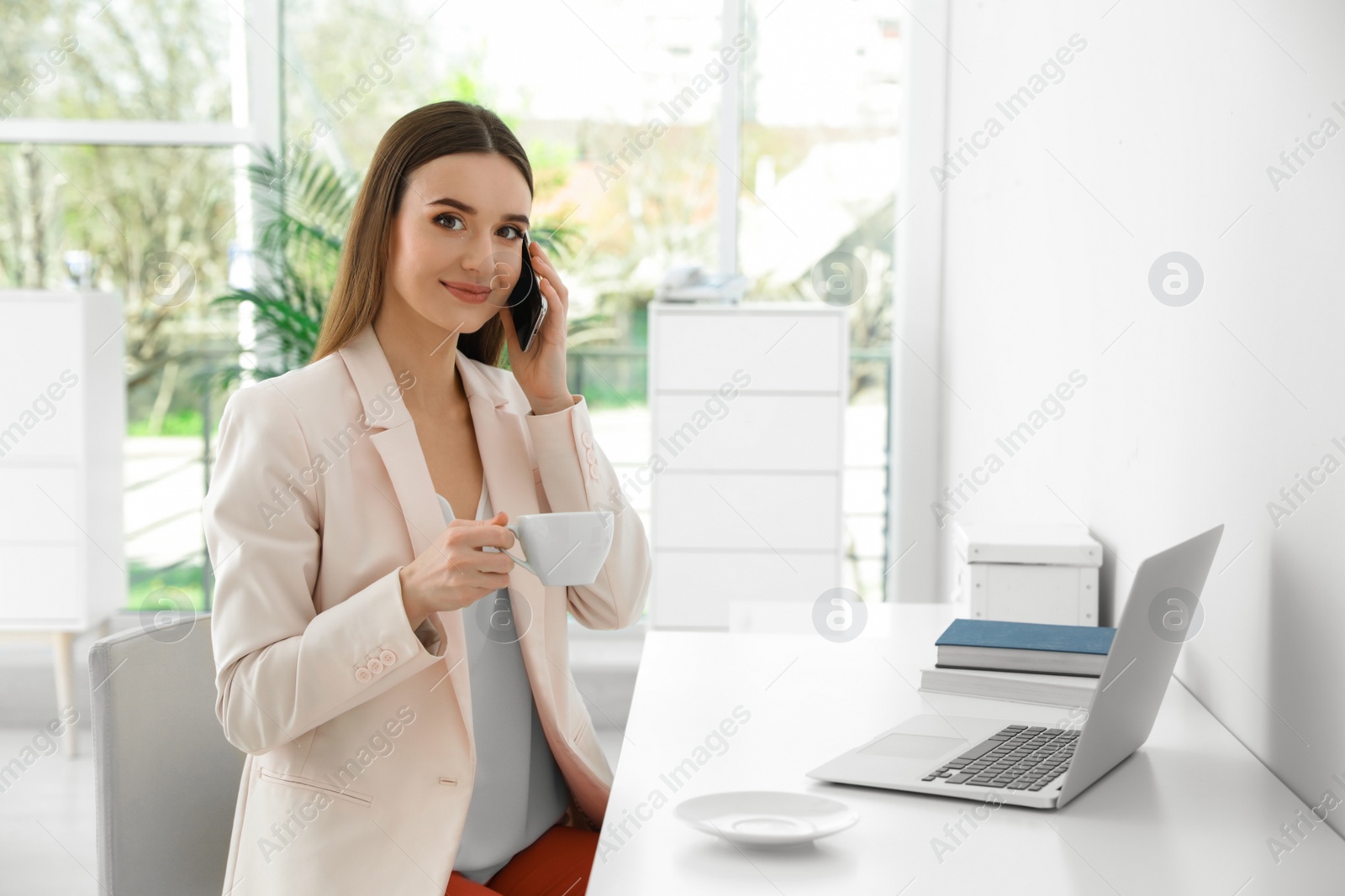 Photo of Young businesswoman talking on phone while using laptop at table in office