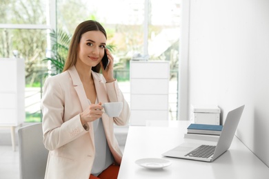 Photo of Young businesswoman talking on phone while using laptop at table in office