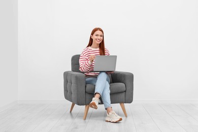 Happy young woman with laptop sitting in armchair near white wall indoors