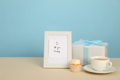 Photo of Happy Father's Day. Tasty cupcake, cup, card with phrase I Love You, Daddy in frame and gift box on beige table. Space for text