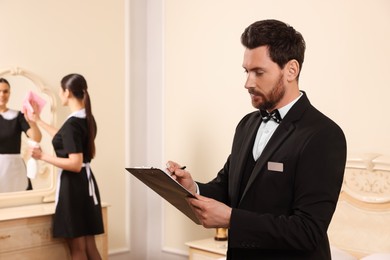 Man wearing suit with clipboard checking maid's work in hotel room, space for text. Professional butler courses