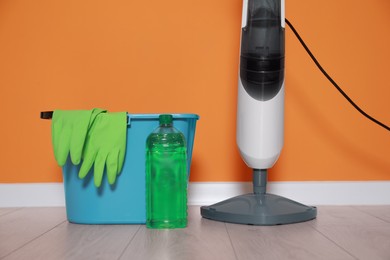 Photo of Modern steam mop, bucket with gloves and bottle of cleaning product on floor near orange wall