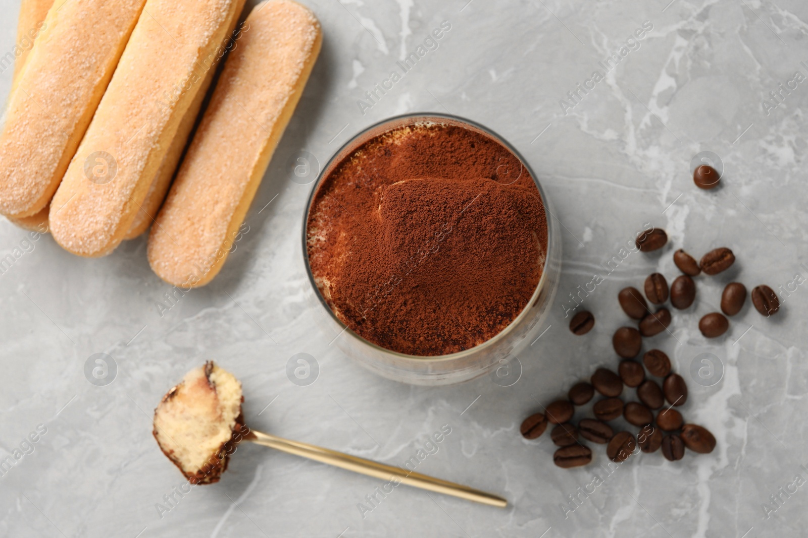 Photo of Tasty tiramisu in glass, coffee beans, spoon and biscuits on light grey table, flat lay