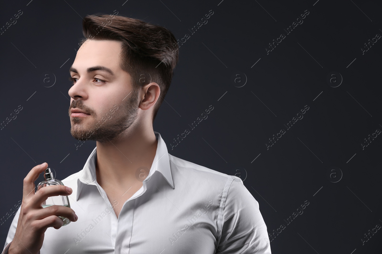 Photo of Handsome man in shirt using perfume on dark background