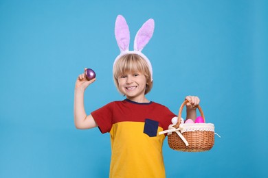 Happy boy in bunny ears headband holding wicker basket with painted Easter eggs on turquoise background