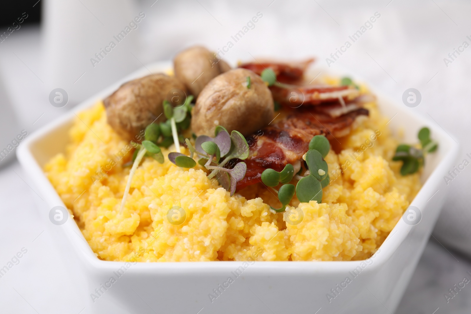 Photo of Cooked cornmeal with bacon, mushrooms and microgreens in bowl on white table, closeup